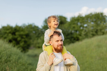 Father with a red beard carrying his toddler on his shoulders while walking outdoors on a sunny day. Warm natural light, joyful family moment and peaceful countryside atmosphere.