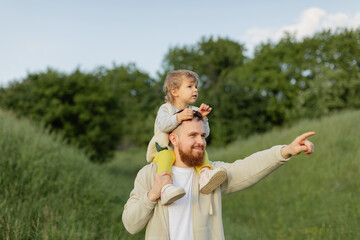 Father with a red beard carrying his toddler on his shoulders while pointing into the distance outdoors. Joyful family moment, warm natural light and summer nature scene.