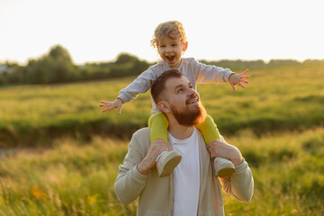 Joyful child spreading arms while riding on her father&rsquo;s shoulders during a sunny golden-hour walk. Warm light, playful energy, and genuine family connection.