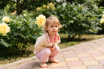 Toddler squatting barefoot in a garden surrounded by blooming yellow peonies. Natural light, calm expression, serene outdoor childhood moment.