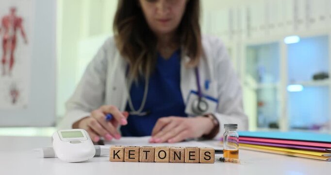 Word Ketones appears on wooden blocks near monitor and sample vial on white desk. Woman clinician checks glucose level recording ketone result