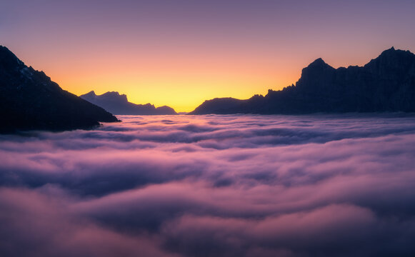 Aerial view of low clouds in the Swiss Alps with mountain silhouettes and vibrant sky and fog-filled valley at sunset. Switzerland. Top view of mountains in low pink clouds and orange sky at twilight