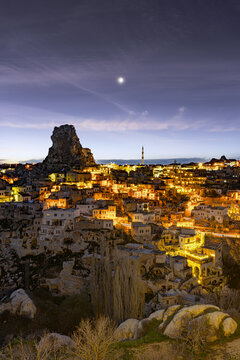 View of a warm, inviting glow emanates from the town nestled among the surreal rock formations under the evening sky, Ortahisar, Cappadocia, Turkey.