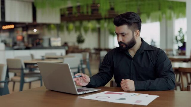 Portrait of focused Indian marketer checks data on papers and laptop screen in coworking. Financial manager studies information about annual commercial activity ar workplace