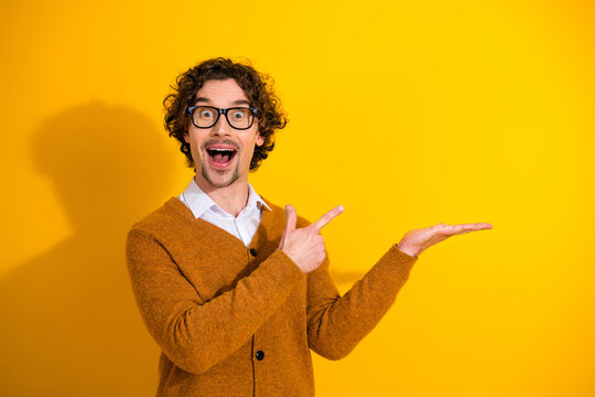 Smiling man with glasses wearing mustard cardigan points to open palm against bright yellow background for advertising lifestyle fashion stock photo