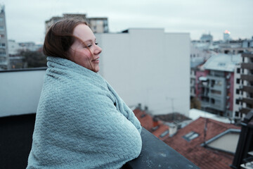 Obraz premium A woman wrapped in a soft blanket stands on a balcony and looks at the urban skyline. The concept expresses reflection and peaceful solitude.