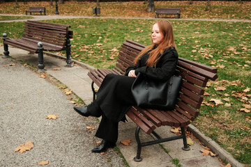 A woman in a black coat sits on a wooden bench in an autumn park, relaxing quietly while enjoying...
