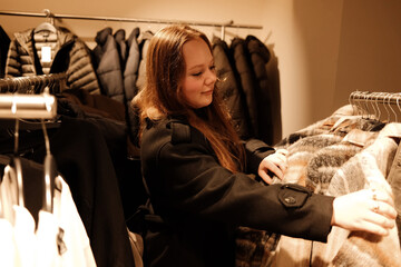 A woman examines a warm patterned coat on a rack, comparing fabrics while shopping for winter...