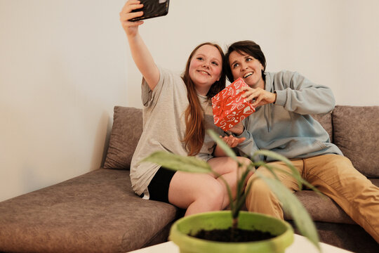 Two friends take a cheerful selfie together while holding a decorated gift box. The concept reflects happiness, celebration and shared friendship moments.