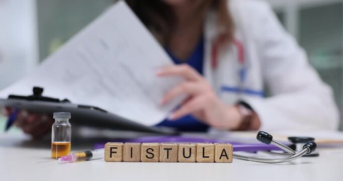Wooden blocks spell word Fistula near vial stethoscope and clipboard on desk. Woman surgeon studies patient record planning treatment for fistula