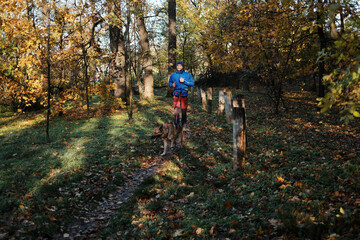 Fototapeta premium Man running with his German Shepherd through a forest trail surrounded by warm autumn colors. The image conveys freedom, nature connection, and quiet movement.