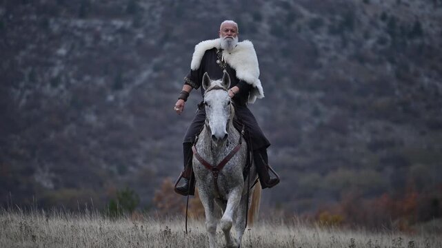 Elderly Viking warrior in fur cloak riding gray horse across open field at sunrise, mountains and distant cross behind him, moody cinematic Norse atmosphere cinematic slow motion 