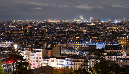 Fototapeta premium Cityscape of Paris, France glows at night seen from Montmartre hill. Panoramic view features illuminated rooftops, streetlights, and urban horizon under cloudy sky in Ile-de-France region