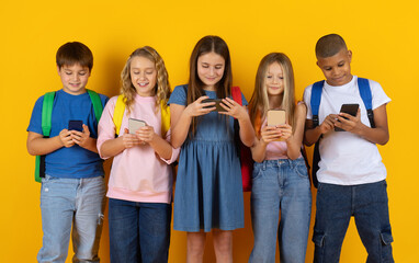 Obraz premium Preteen pupils standing together while looking at smartphones in bright studio background. A modern group of children engaged with technology in a school lifestyle scene