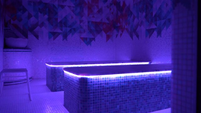 Modern illuminated hammam spa room with glowing mosaic massage tables, vibrant geometric tile walls, and atmospheric purple LED lighting creating a relaxing wellness interior