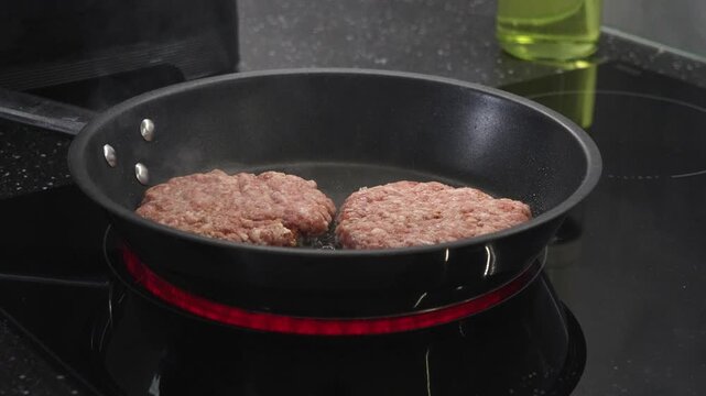 Two raw beef burger patties sizzling actively in a black non-stick frying pan on a glowing electric induction stovetop, representing easy home cooking