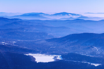 Fototapeta premium Panoramic view of mountain ridges near Sheregesh, Russia, covered in soft blue haze. Peaks fades into distance, creating depth effect under winter sky. Scenic landscape with frozen lake, reservoir.
