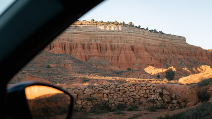 A scenic view of layered red cliffs captured from a car window. The landscape shows warm desert tones, rock formations and a dramatic sky, creating a natural travel and adventure mood.