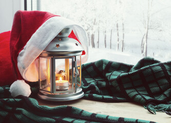 Festive Christmas and New Year still life - lantern with candle and Santa hat on the windowsill and winter forest outdoor. Concept of Christmas and New Year. Christmas and New year background
