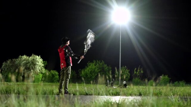 Night race marshal waving checkered flag under bright floodlight, blurred kart streaks past finish on wet