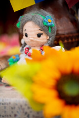 Close-up of a handmade crocheted doll with grey braids and flower accessories, partially obscured by a bright orange sunflower in the foreground