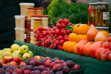Fresh vegetables and fruits displayed at a local market, including radishes, tomatoes, apples, and plums, with jars of homemade preserves in the background
