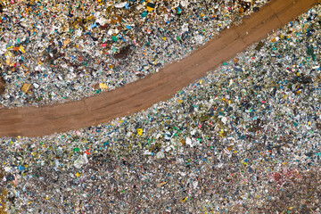 Aerial view of a landfill with a winding dirt road cutting through large areas of mixed waste and plastic debris.