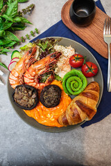 A gourmet breakfast plate with shrimp, croissant, avocado rose, scrambled eggs, salad, and mushrooms.