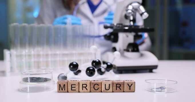 Word Mercury of wooden blocks against metal samples and molecular structure model on desk. Woman scientist checks goggles before tests start in lab