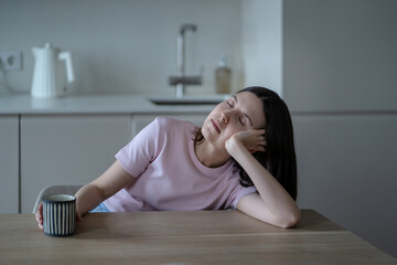 Exhausted sleepy woman sitting at kitchen table with coffee, leaning head on hand, closing eyes, dozing off early in morning. Sleepiness, morning fatigue of drowsy female after sleepless unrest night