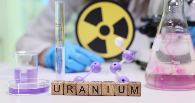 Word Uranium on wooden blocks near purple liquids and molecular structure in lab. Woman chemist researches radioactive metal ore by radiation symbol