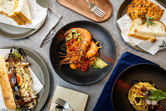 Grilled prawns over pasta with tomato sauce and zucchini, surrounded by gourmet sides like chicken, bread, and salad. Elegant dining setup.