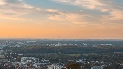 Naklejka premium Panoramic view of a sprawling urban landscape at sunset, showcasing buildings, greenery, and distant industrial structures under a colorful sky, representing city life and nature