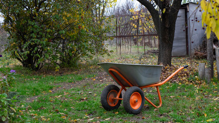 Double-Wheeled Orange and Gray Wheelbarrow Sitting on a Grassy Path in an Overgrown Autumn Garden