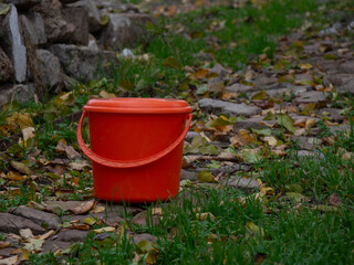 Bright Orange-Red Plastic Bucket with a Lid Sitting on a Stone Path Covered with Autumn Fallen Leaves
