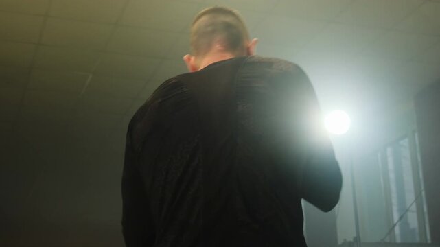 Boxer simulates real sparring in spacious dimly lit hall. Man shadowboxes and throws punches professionally standing in posture repositioning body