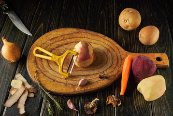 Fresh vegetables are being prepared on a wooden cutting board. A potato is being peeled with a yellow peeler, surrounded by other colorful ingredients like carrots and onions