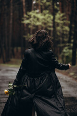 Naklejka premium Woman in a black coat walks down a forest path holding a yellow rose.