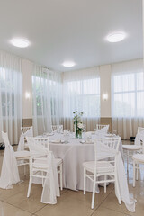 Elegant wedding reception setup with white decor and floral centerpiece in a sunlit room.