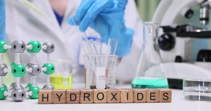 Young woman chemist carefully adds reagent to liquid in test tube slow motion. Miniature wooden cubes on laboratory table form term Hydroxides