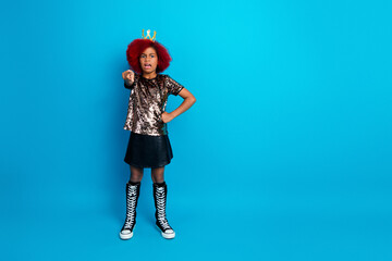 Young girl with red hair crown sequined top black skirt and tall boots points at camera with confidence against a bright blue studio background