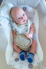 A baby relaxes in a soft seat at home. The baby has blue eyes and is wearing a light shirt and colorful socks. The room has a calm atmosphere with gentle lighting