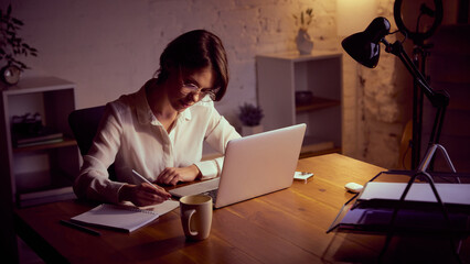 Woman studying at laptop in cozy evening home office. Concept of online education, remote work...