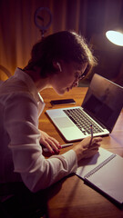 Woman writing notes near laptop with wireless earbuds during study session. Concept of online education, hybrid learning routine, digital course materials, and focused study workflow.