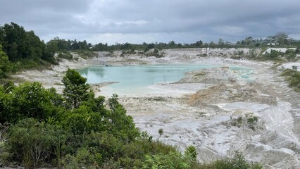 A serene view of a light blue-green lake surrounded by white and brown barren land with scattered green vegetation under an overcast sky.