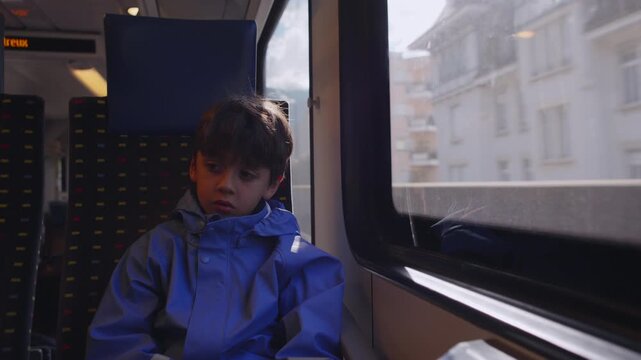 Young boy leaning toward train window with subdued expression while traveling through town, creating quiet contemplative mood during slow regional rail journey