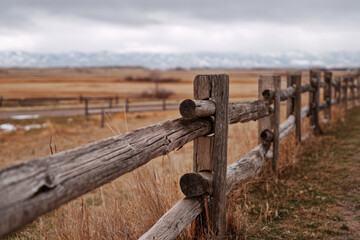 A weathered wooden fence lines the path to a rustic barn on a ranch, surrounded by open fields and distant mountains under gray skies, capturing the peaceful rural atmosphere