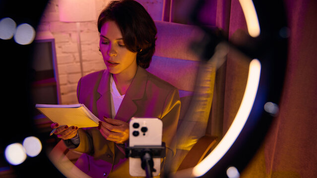 Woman reading notes while filming educational video on phone. Concept of structured content creation, expert digital storytelling, and calm professional communication.