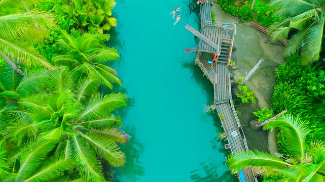 Aerial top-down view of two young women swimming in the vibrant turquoise Maasin River, surrounded by lush tropical palm trees and wooden docks in Siargao, Philippines.
