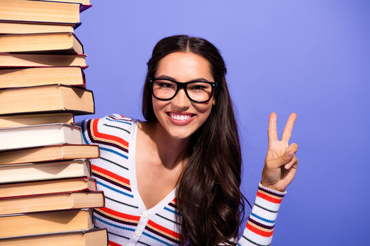 Young woman with glasses smiles beside a tall stack of books and shows a peace sign against a blue background